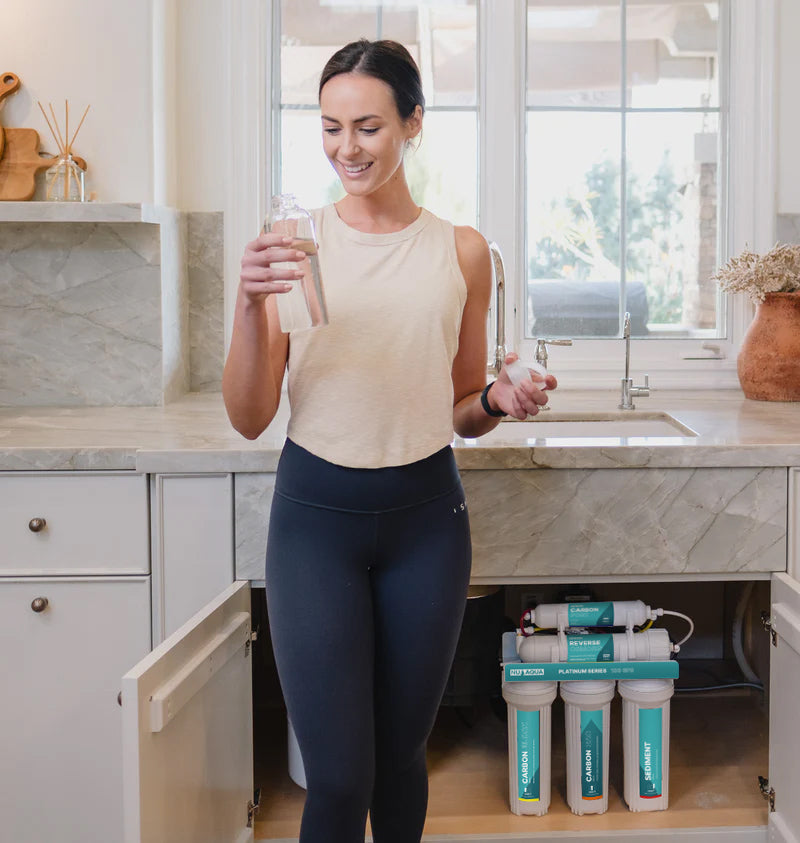 Woman Smiling And Holding Glass Of Reverse Osmosis Filtered Water In Front Of Kitchen Sink